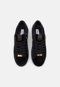 Černé semišové tenisky s plochými tkaničkami, zaoblenou špičkou a zlatou a černou logovou akcentací na boku. Texturovaný svršek s kontrastními detaily.