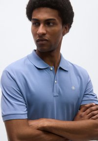 Ανοιχτό μπλε πόλο μπλούζα από βαμβακερό ύφασμα, με τρία κουμπιά και μικρό κεντημένο λογότυπο στο αριστερό στήθος. Κοντά μανίκια.