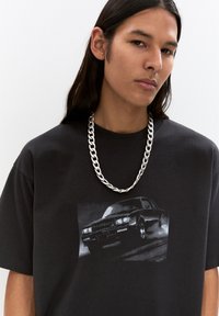 Black cotton t-shirt featuring a graphic of a classic car in grey, complemented by a thick silver chain necklace worn around the neck.