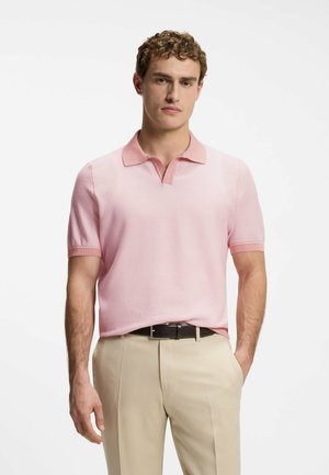 Uomo che indossa una polo a maniche corte rosa chiaro infilata in pantaloni beige con cintura nera, in piedi davanti a uno sfondo bianco uniforme.