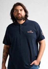 Marinblå polo med klassisk krage, två knäppningar och broderade lappar på ärmarna. Mjuk tyg och avslappnad passform.