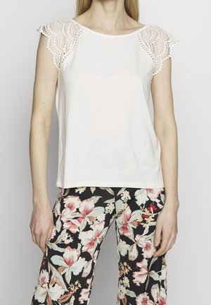 Weißes Oberteil mit Spitzen-Details an den Schultern, abgerundeter Ausschnitt, kombiniert mit locker sitzender Hose mit floralem Muster in Schwarz, Pink und Grün.