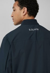 Marineblauwe jas met een zachte textuur, voorzien van een staande kraag en het logo "S.OLIVER" geborduurd op de achterkant.