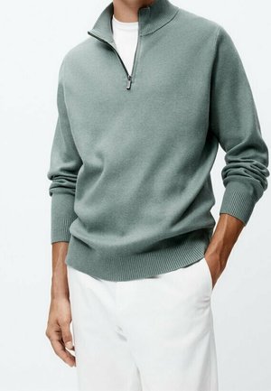 Homme portant un pull à col zippé sage vert et un pantalon blanc, une main dans la poche et l'autre détendue le long du corps.