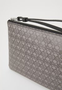 Bolso de mano gris con textura, patrón en relieve, cremallera negra y correa de muñeca de cuero; diseño elegante sin hardware adicional.