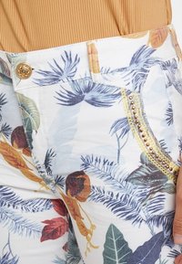 Bílé kalhoty s květinovým vzorem, které mají zlatý knoflík, dekorativní korálky na kapse a lehkou látku s hladkou texturou.