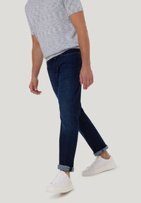 Man in opgerolde donkerblauwe jeans, lichtgrijs kortemouwen shirt en witte sneakers, staande tegen een effen achtergrond.