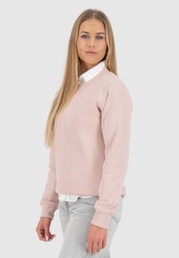 Felpa rosa chiaro con scollo rotondo, polsini e orlo ribattuti, indossata sopra una camicia bianca con colletto, abbinata a jeans grigio chiaro.