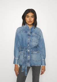 Denim srajca z dolgimi rokavi v svetlo modri barvi, z pasom, gumbi spredaj in dvema žepoma na prsih. Ima srednje težko tkanino in sproščen kroj.