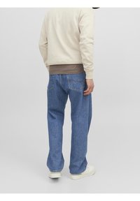 Ljusbeige sweatshirt över en brun topp, kombinerad med löst sittande blå denimjeans. Vita skor fullbordar outfiten. Enfärgad bakgrund.