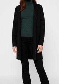 Svart lång cardigan i mjukt tyg, öppen frontdesign, kombinerad med en grön polotröja och svarta tajta jeans, minimala detaljer.