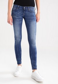 Jeans in denim blu slim-fit con un effetto sbiadito, cinque tasche, chiusura a bottone e una leggera elasticità, abbinati a sneakers bianche.