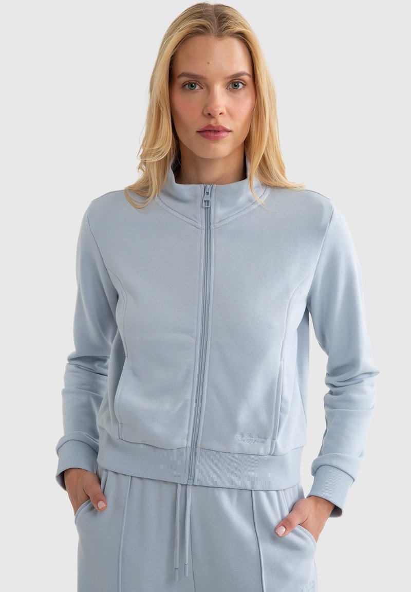 Gaiši zils rāvējslēdzēja džemperis, izgatavots no mīkstas auduma, ar augstu apkakli, ribotu piedurknēm un pieguļošu dizainu ar smalkām šūšanas detaļām.