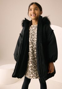 Black puffer coat with fur-trimmed hood, oversized shape, zip closure, worn over a beige leopard print dress, paired with black leggings.