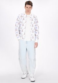 Lichte bomberjack met bloemenprint, witte basis, gecombineerd met lichtblauwe cargobroek. Model draagt witte sneakers en staat tegen een effen achtergrond.