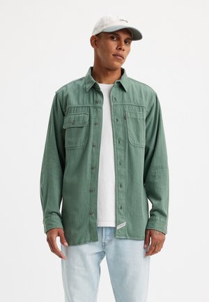 Green button-up shirt made of soft fabric, features two chest pockets, metal buttons, layered over a white t-shirt, worn with light denim jeans.