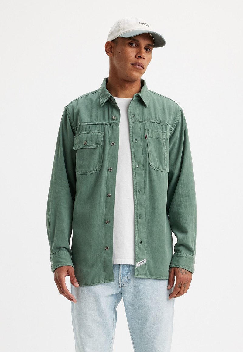Camisa verde de corte recto hecha de tela suave, con dos bolsillos en el pecho, botones metálicos, superpuesta sobre una camiseta blanca, llevada con jeans de mezclilla claros.