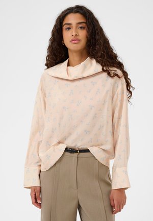 Žena s dlhými kučeravými vlasmi, oblečená v svetloružovej kvetinovej blúzke so širokým golierom a béžových vysokých nohaviciach s čiernym opaskom.