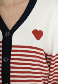 Cardigan à rayures blanches et rouges avec des bords bleu marine, présenté avec des boutons noirs et un petit détail en forme de cœur rouge en haut à gauche.