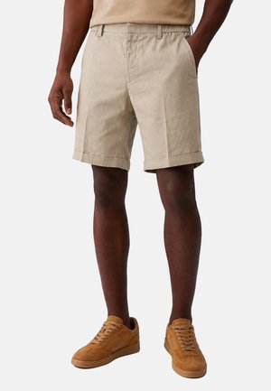 Uomo che indossa pantaloncini beige al ginocchio e sneakers in camoscio color marrone chiaro, in piedi con una mano in tasca su sfondo neutro.