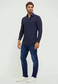 Camisa azul marinho de botão, com mangas longas, tecido suave, corte ajustado, combinada com jeans de ganga escura e ténis brancos.