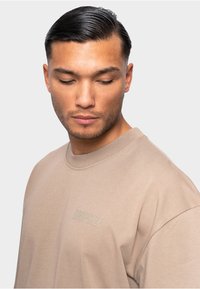 T-shirt en coton beige avec un col rond et des manches courtes. Présente un logo en relief "DROPSIZE" sur la poitrine gauche, texture lisse.