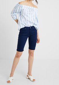 Blusa de rayas azul claro y blanca con escote off-the-shoulder y mangas abullonadas, combinada con pantalones cortos de mezclilla azul oscuro y sandalias blancas.