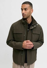 Olivno zelena jakna iz gladkega, vodoodpornega materiala; ima dva žepka na prsih, visok ovratnik in detajle s pritisnimi gumbi.