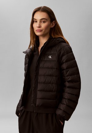 Frau mit glattem braunem Haar, die eine schwarze gesteppte Jacke mit cK-Logo trägt, steht vor einem einfarbigen grauen Hintergrund, die Hände in den Taschen.