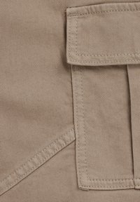 Beige tyg med ett diagonalt vävmönster, med en stor klaffficka och välgjorda sömmar längs kanterna för hållbarhet.