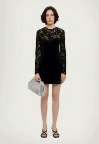 Vestido de encaje negro de manga larga, con cuerpo ajustado y falda acampanada. Combinado con un clutch plateado y sandalias negras con tiras. Detalles suaves y texturizados.