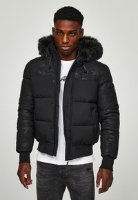 Chaqueta de plumón negra con un patrón de camuflaje texturizado, capucha forrada de piel, cierre con cremallera y puños acanalados. Llevada sobre una camiseta blanca.