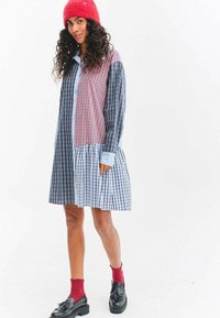 Žena oblečená v šatové košili s kostkovaným vzorem, červené pletené čepici, červených ponožkách a černých mokasínách, stojící před bílým pozadím.