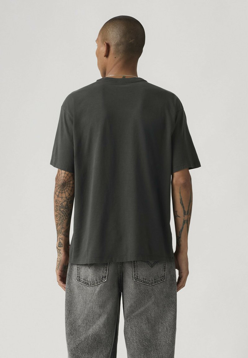 Homme vu de dos portant un t-shirt ample sombre uni et un jean gris délavé, avec des tatouages visibles sur les deux bras.