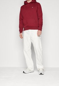 Sudadera con capucha color marrón burdeos y bolsillo frontal, combinada con pantalones blancos de corte holgado. Completada con zapatillas de running grises y negras.