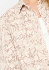Light beige button-up shirt with a subtle snake print pattern, featuring a collar and a patch pocket on the left side. Soft texture.
