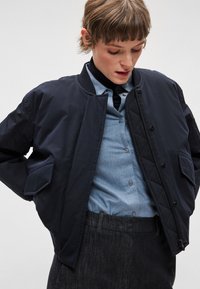 Tumši zila jūras virsnieka jaka ar šūtu dizainu, kas aprīkota ar ribotu apkakli, priekšējām pogām un sānu kabatām, valkāta pār gaiši zilu kreklu.