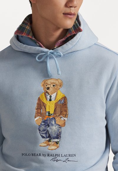 Φωτεινό μπλε φούτερ με γραφικό άγριου θηρίου ντυμένο με καρό σακάκι, κίτρινο πουλόβερ και τζιν. Συνοδεύεται από λογότυπο κάτω από το σχέδιο.