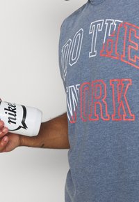 Camiseta sin mangas gris con gran texto blanco y rojo que dice "HACE EL TRABAJO". Botella de agua blanca de Nike con logo negro sostenida en la mano.