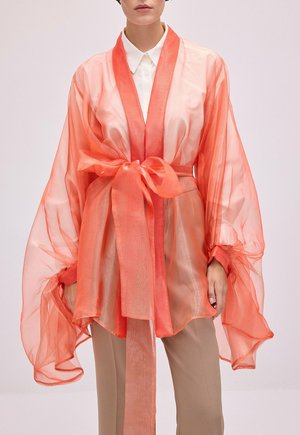 Femme portant une veste style kimono orange corail transparente avec de larges manches et un nœud noué à la taille, par-dessus une chemise blanche et un pantalon beige.