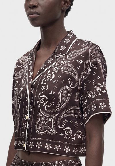 Gros plan d'une personne portant une chemise marron foncé à manches courtes boutonnée avec des motifs paisley et floraux blancs ainsi qu'un liseré blanc le long des bords.