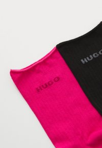 To par sokker, en pink og en sort, med ribbet tekstur. Hver har mærket "HUGO" trykt i tilsvarende kontrastfarver.