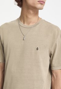 Beige kortærmet t-shirt med rund hals, præget af et lille mørkt logo på venstre bryst. Glat, afslappet stoftekstur.