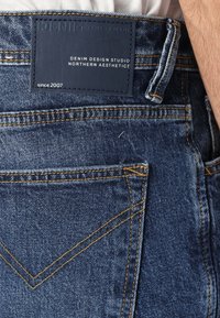 Un jean en denim bleu foncé présente une surface texturée, des coutures jaunes et une étiquette en cuir de marque sur la taille au dos.