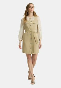 Abito beige con un design aderente, chiusura a bottoni, due tasche sul petto e una cintura in vita. Abbinato a una blusa trasparente a maniche lunghe.