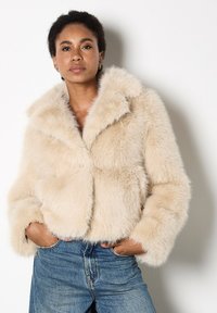 Faux fur cropped jasje in zacht beige met een brede kraag, pluche textuur en verlaagde schouders, gecombineerd met high-waisted denim jeans.