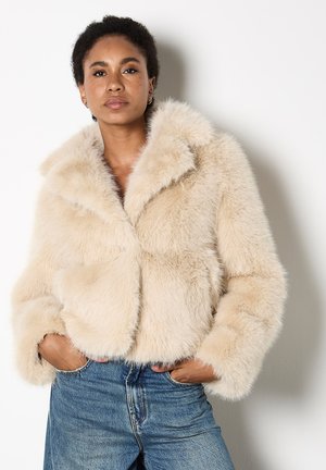 Faux fur cropped jasje in zacht beige met een brede kraag, pluche textuur en verlaagde schouders, gecombineerd met high-waisted denim jeans.