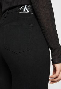 Jeans de ganga pretos com uma cintura alta, apresentando um logótipo da Calvin Klein e um bolso traseiro com um contorno costurado. Textura suave.