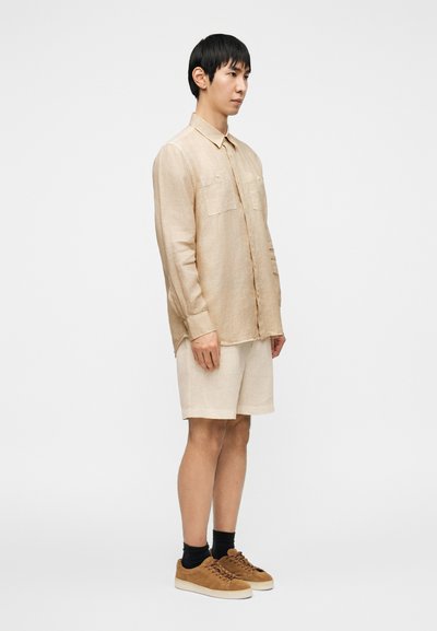 Ανοιχτό μπεζ λινό πουκάμισο με δύο τσέπες στο στήθος, μακριά μανίκια και άνετη εφαρμογή, σετ με matching σορτς και καφέ σουέτ αθλητικά παπούτσια.
