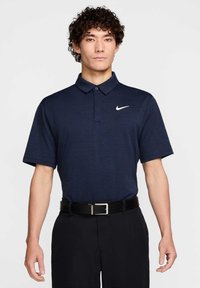 Tamsiai mėlyna polo marškinėliai, pagaminti iš tekstūruotos medžiagos, su dviem mygtukais ir baltu Nike logotipu kairiajame krūtinės pusėje. Juodas diržas su sidabriniu sagtimi.
